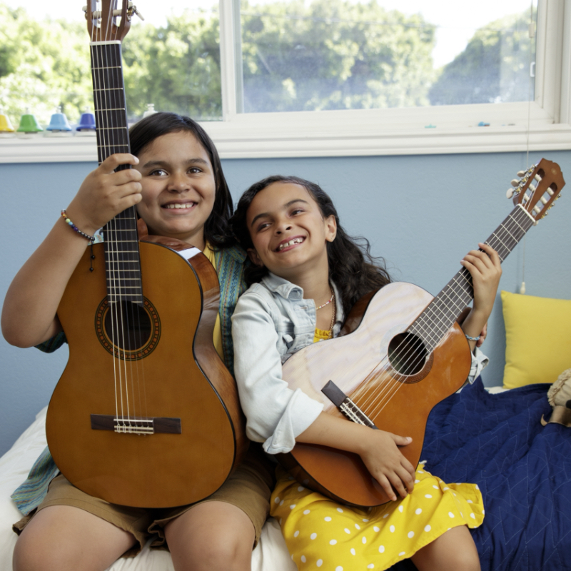 kitare-blog-1 Two girls sitting close together with acoustic guitars, smiling at the camera.