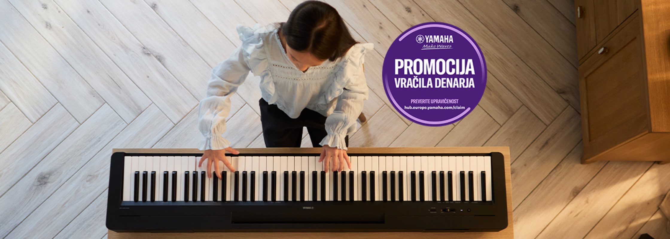 Top-down view of a person playing a digital piano on a light wood floor, with a purple Yamaha promo sticker nearby.